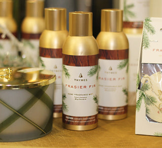 Thymes Frasier Fir home fragrance mist bottles with gold caps and brown-and-white labels, displayed on a tabletop with a candle and gift box nearby.