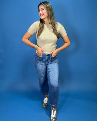A woman posing in a beige short-sleeve knit top, blue cropped jeans, and white sneakers against a solid blue studio backdrop.
