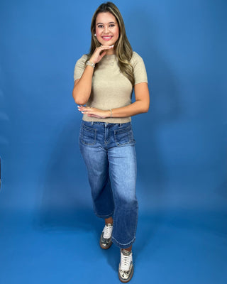 A woman posing in a beige short-sleeve knit top, blue cropped jeans, and white sneakers against a solid blue studio backdrop.