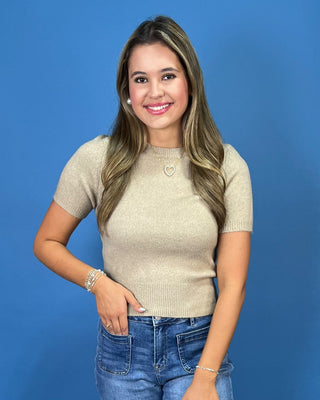 A woman posing in a beige short-sleeve knit top, blue cropped jeans, and white sneakers against a solid blue studio backdrop.