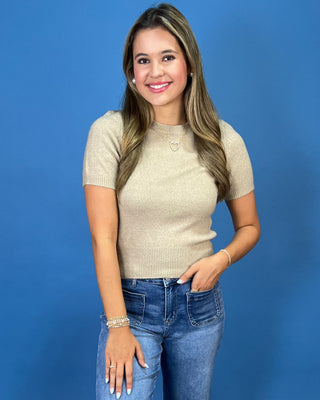 A woman posing in a beige short-sleeve knit top, blue cropped jeans, and white sneakers against a solid blue studio backdrop.