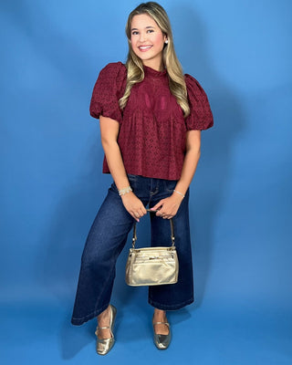 A woman modeling a burgundy eyelet puff-sleeve blouse with a high mock neck, paired with blue jeans