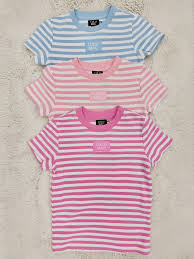 Three short-sleeve striped t-shirts stacked vertically on a textured beige background. The top t-shirt is light blue and white striped, the middle is pink and white striped, and the bottom is pink and magenta striped.