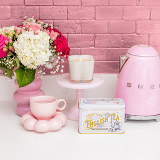 Pink tea set with a teapot, cup, and English tea box against a pink brick wall.