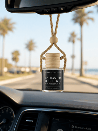A car air diffuser bottle labeled 'Enchanted Rouge' hanging from a rope harness under the rearview mirror inside a car,