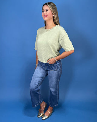 A woman modeling a light sage green short-sleeve knit top and blue wide-leg jeans against a solid blue studio backdrop.
