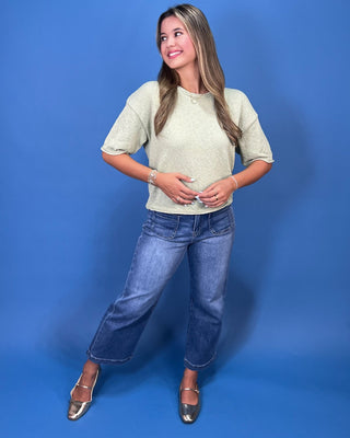 A woman posing in a light sage green short-sleeve knit top, blue cropped jeans, and metallic silver shoes against a solid b