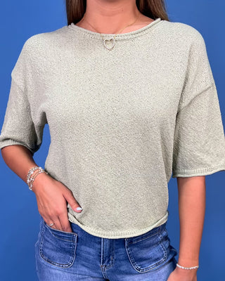 A woman wearing a light sage green knit short-sleeve crop top with a small silver heart detail at the neckline, paired with b