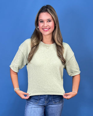 A smiling young woman with long highlighted brown hair stands against a solid blue studio background, wearing a short-sleev