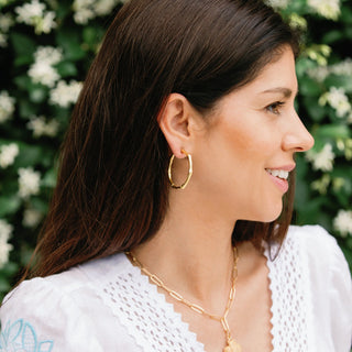 Woman wearing gold hoop earrings and a necklace with a blurred green and white floral background