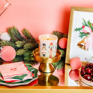Decorative Christmas setting with a lit candle, 'JOY' napkins, and festive decor on a pink background.