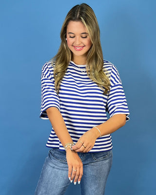 A woman wearing a blue and white horizontally striped short-sleeve top and blue jeans, standing against a solid blue stu