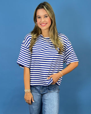 A woman wearing a blue and white horizontally striped short-sleeve t-shirt and light wash blue jeans, standing against a so