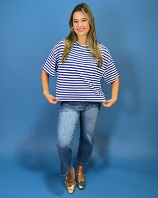 blue and white horizontally striped short-sleeve t-shirt and blue jeans, standing against a solid blue studio background.