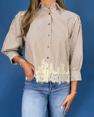 A woman modeling a beige and white vertically striped blouse with a cream lace hem, paired with blue jeans and metallic sand