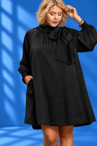 A woman wearing a black long-sleeve tunic dress with a large bow at the neck and side pockets