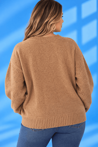 A woman shown from the back wearing a camel brown knit sweater with textured eyelet details on the sleeves, paired with blue