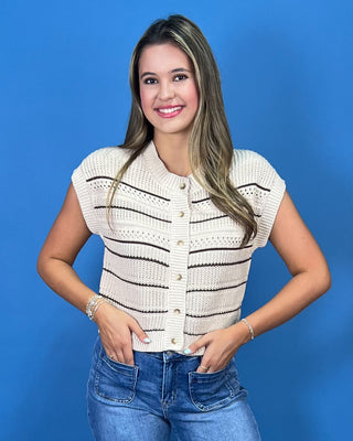 A woman modeling a cream knit cropped vest with thin black horizontal stripes, paired with blue jeans and metallic flats