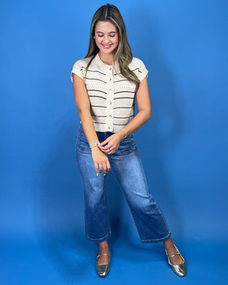 A woman modeling a cream knit cropped vest with thin black horizontal stripes, paired with blue jeans and metallic flats