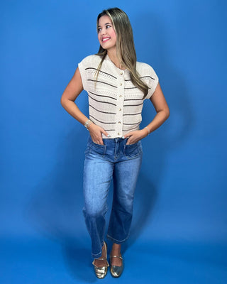 A woman modeling a cream knit cropped vest with thin black horizontal stripes, paired with blue jeans and metallic flats