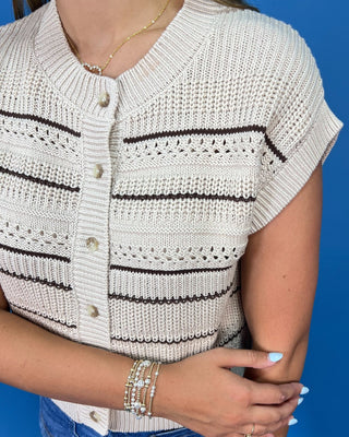 A woman modeling a cream knit cropped vest with thin black horizontal stripes, paired with blue jeans and metallic flats