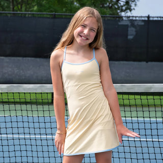 A young girl with blonde hair stands on a tennis court wearing a pale yellow tennis dress with light blue trim. The dress has