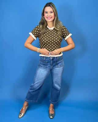 A woman modeling a brown short-sleeve top with white polka dots and white collar/cuff trim, paired with blue jeans and metal