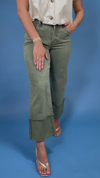 A model wearing a pair of olive green wide-leg cropped jeans with rolled cuffs, paired with a white blouse