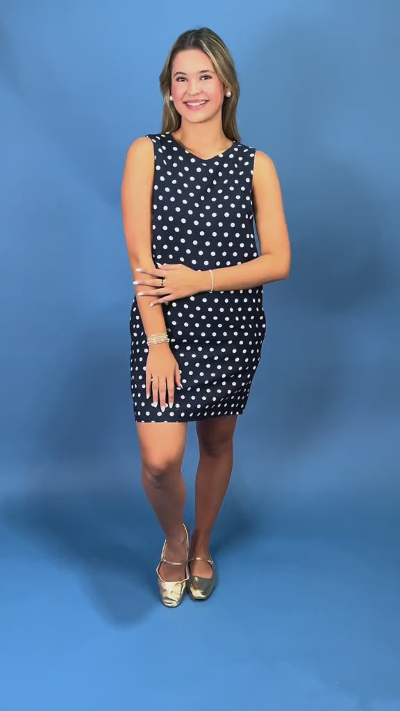 A woman modeling a sleeveless navy blue and white polka dot dress against a solid blue studio background.
