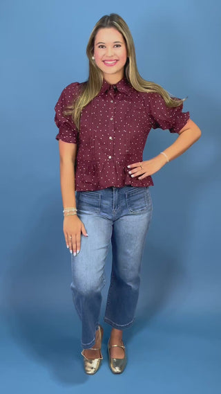 A woman modeling a short-sleeve button-up blouse with puff sleeves and a white polka-dot pattern on a burgundy base, paired with blue jeans, against a solid blue studio background.