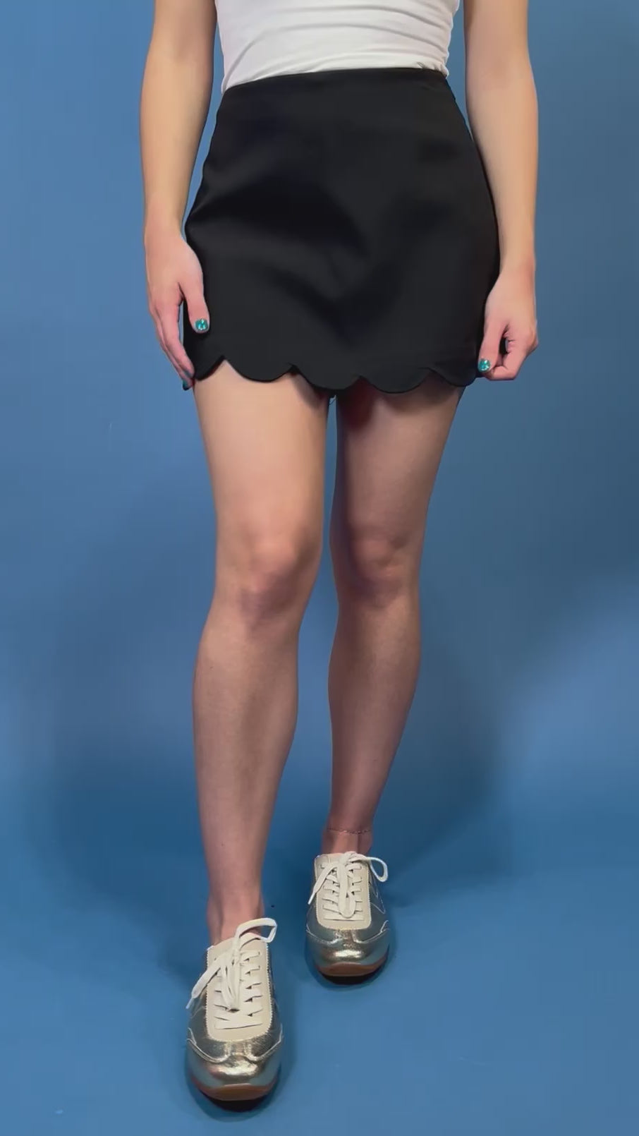 A person modeling a black mini skirt with a scalloped hem, paired with a white top and white sneakers