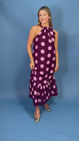 A woman modeling a sleeveless halter-neck maxi dress with a dark purple base and large light pink polka dots