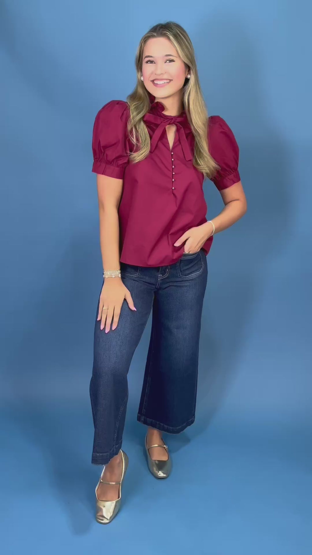 A woman modeling a short-sleeve burgundy blouse with puff sleeves, a keyhole tie neckline, and dark blue wide-leg jeans