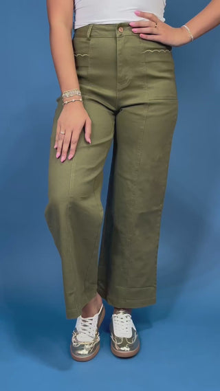 A person wearing olive green wide-leg cropped pants with decorative zigzag stitching on the front pockets, paired with a white top and white sneakers