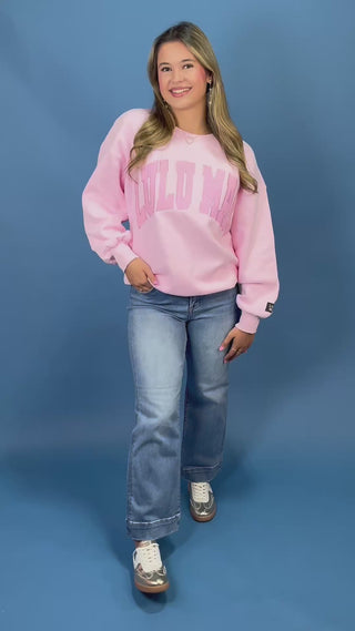 A woman modeling a light pink crewneck sweatshirt with 'LULU MAC printed in darker pink across the chest, paired with blue jeans and metallic sneaker