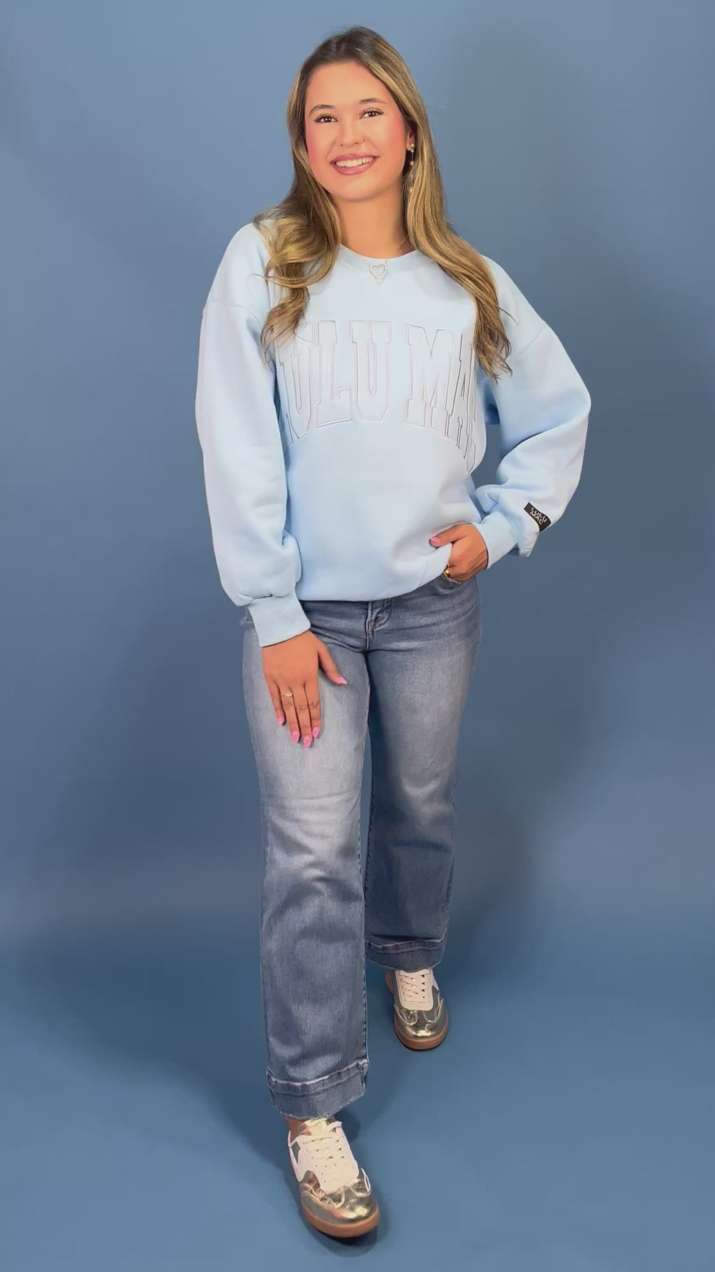 A woman modeling a light blue sweatshirt with 'LULU MAC' printed across the chest, paired with blue jeans and gold sneaker