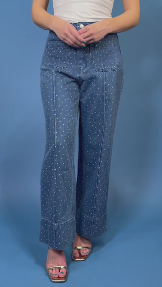 A person wearing blue denim jeans with a white polka-dot pattern, paired with a white top and metallic sandals