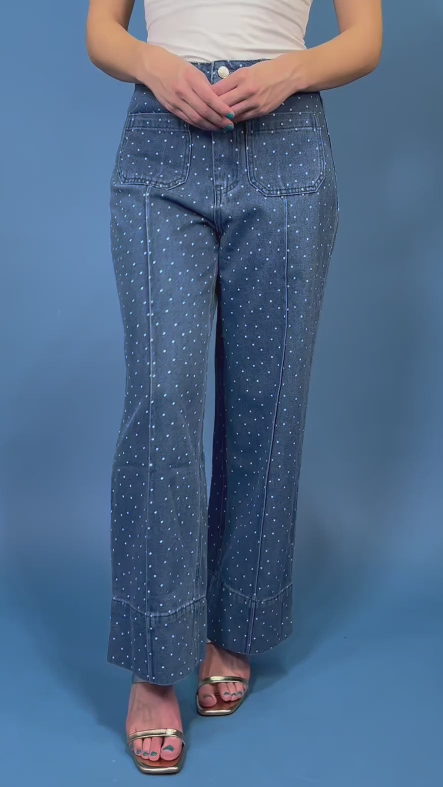 A person wearing blue denim jeans with a white polka-dot pattern, paired with a white top and metallic sandals