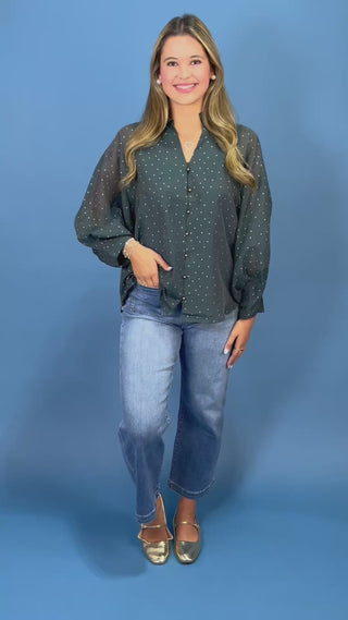 A woman modeling a dark green sheer long-sleeve blouse with small white polka dots, paired with blue jeans and metallic flats, standing against a solid blue studio backdrop.