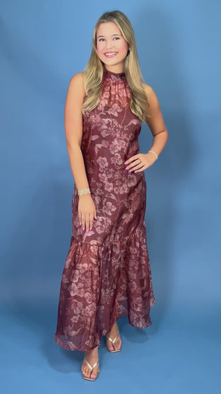 A woman modeling a sleeveless, high-neck, floor-length halter dress with a dusty rose floral pattern on a brownish-rose base