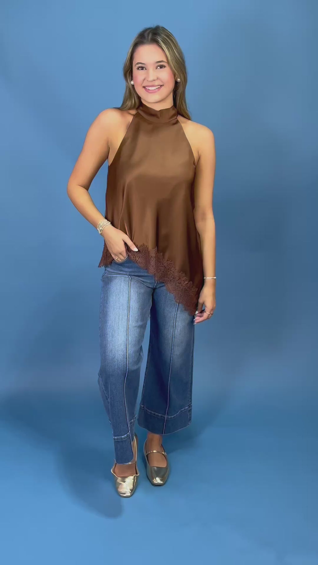 A woman modeling a brown satin halter top with a lace hem detail, paired with blue jeans