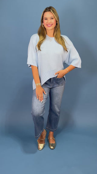 A woman wearing a light blue short-sleeve knit top and blue jeans, standing against a solid blue studio background.