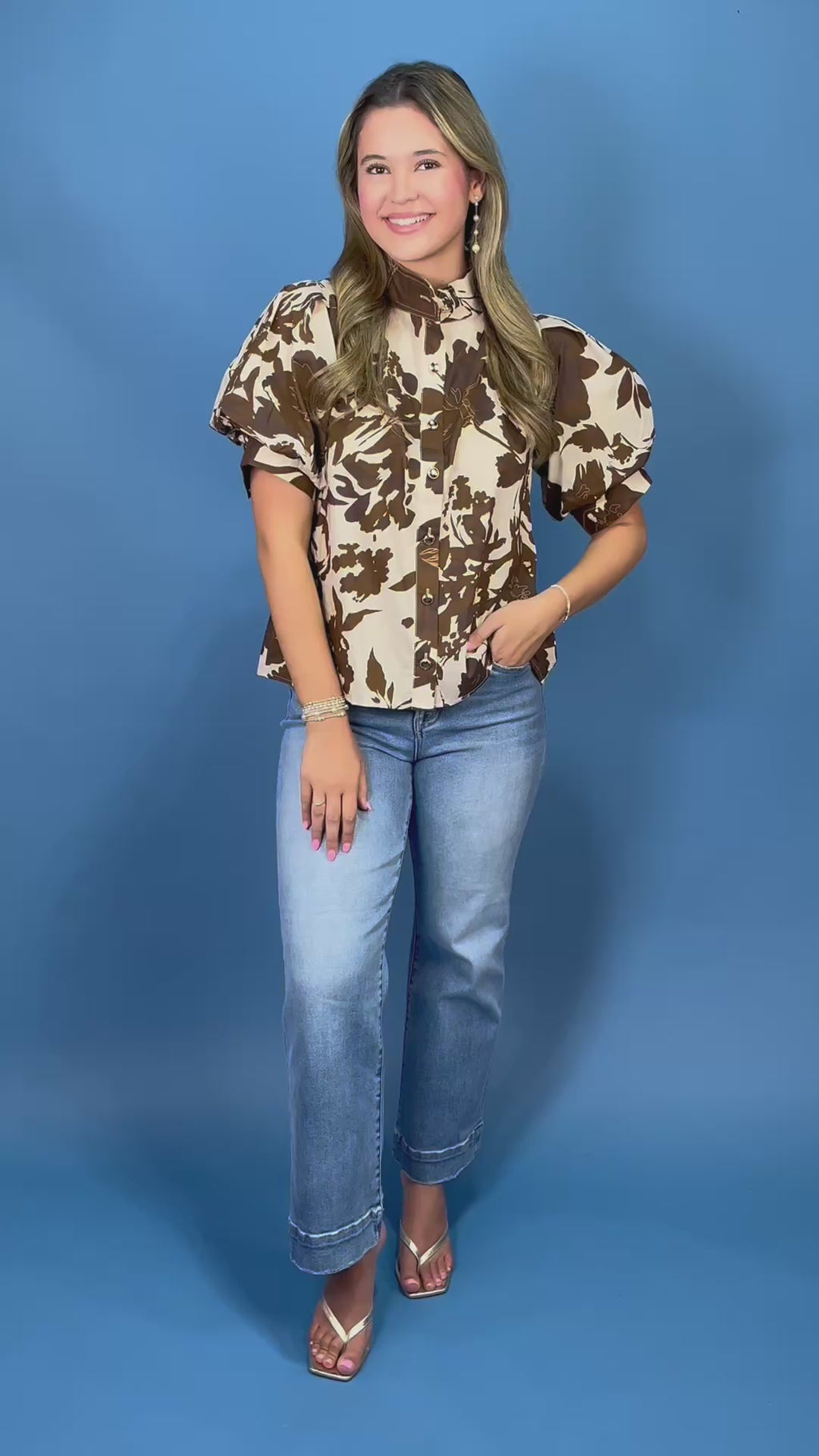 A woman modeling a short-sleeve button-up blouse with a brown floral print on a cream background, paired with blue jeans