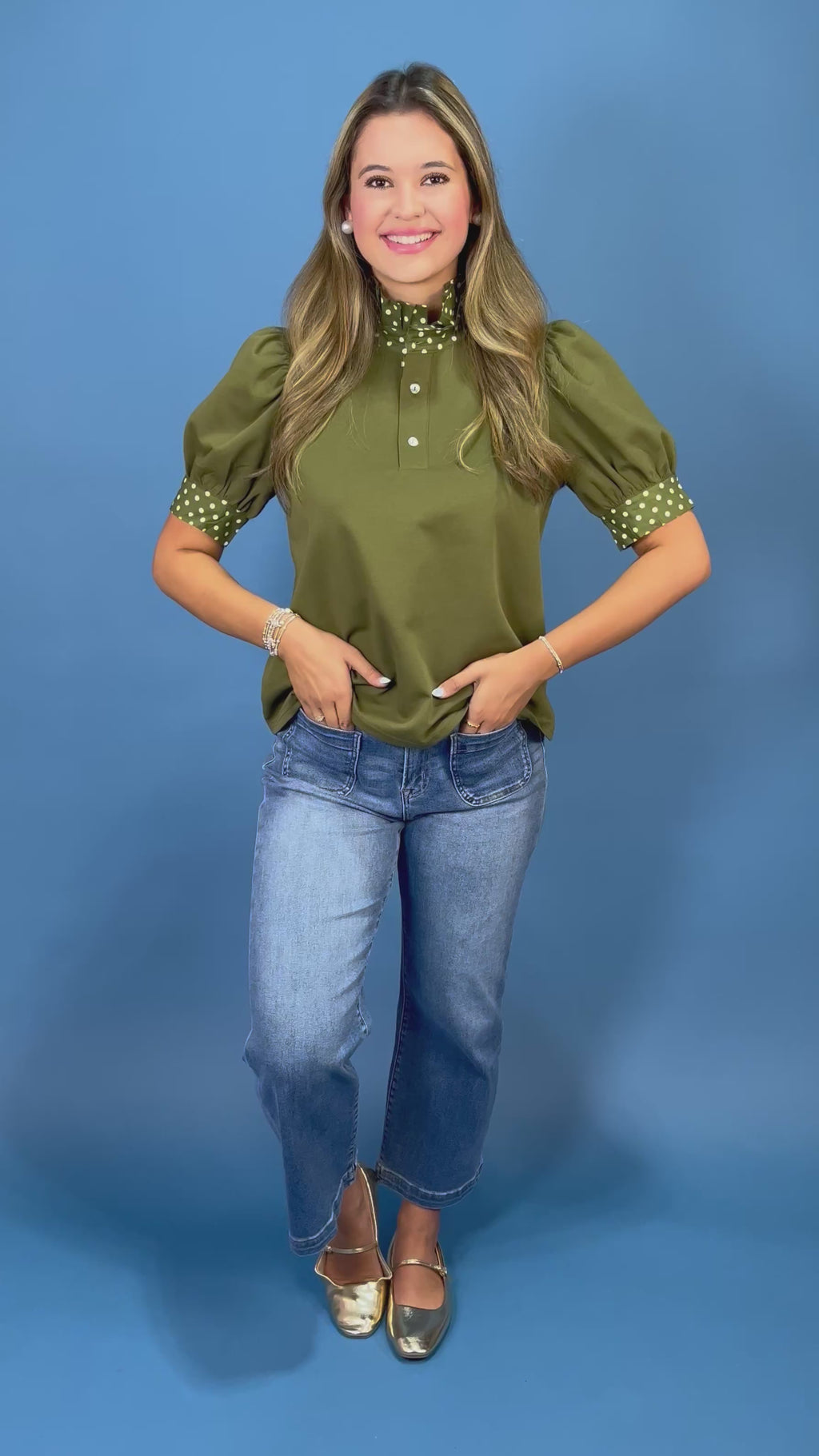 A woman modeling an olive green short-sleeve blouse with white polka-dot trim on the collar and cuffs, paired with blue jeans, against a solid blue studio background.