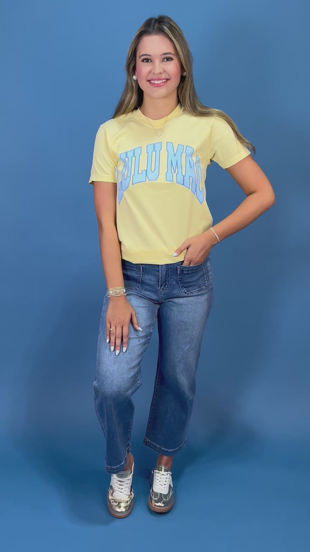 A woman posing in a light yellow graphic t-shirt with blue lettering, paired with blue jeans