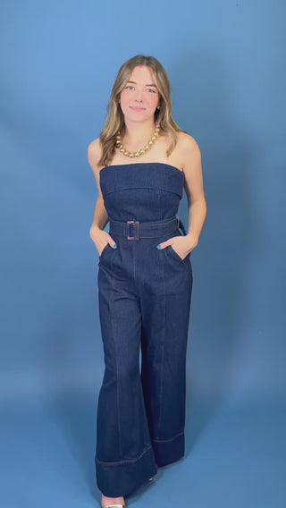 A woman modeling a dark blue denim strapless jumpsuit with wide legs and a matching belt