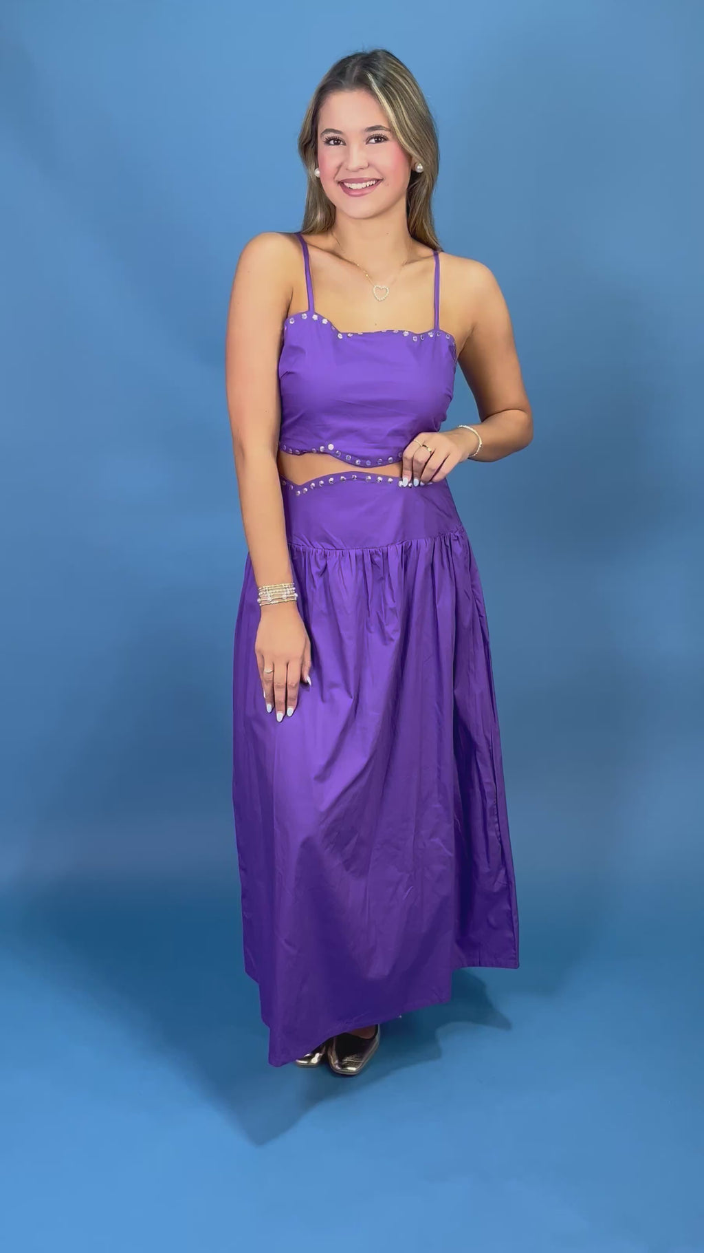 A woman modeling a purple two-piece maxi dress with a crop top and flared skirt, featuring silver rhinestone trim, set against a solid blue studio background.
