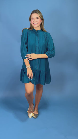 A woman modeling a teal blue mini dress with three-quarter balloon sleeves, a high mock neck, and a ruffled hem