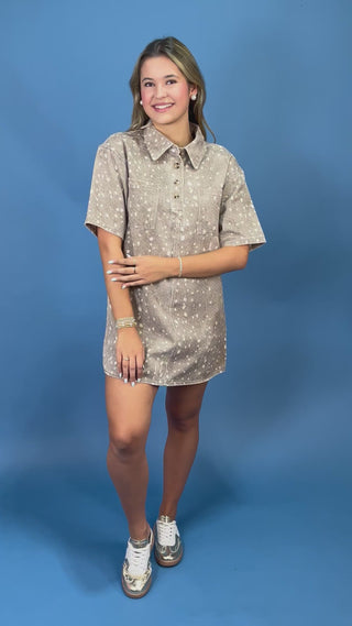 A young woman modeling a short-sleeve button-up shirt dress with a beige and white speckled pattern, standing against a solid blue studio background.