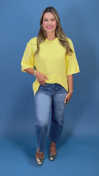 A woman wearing a bright yellow short-sleeve knit top and blue jeans, standing against a solid blue studio background.
